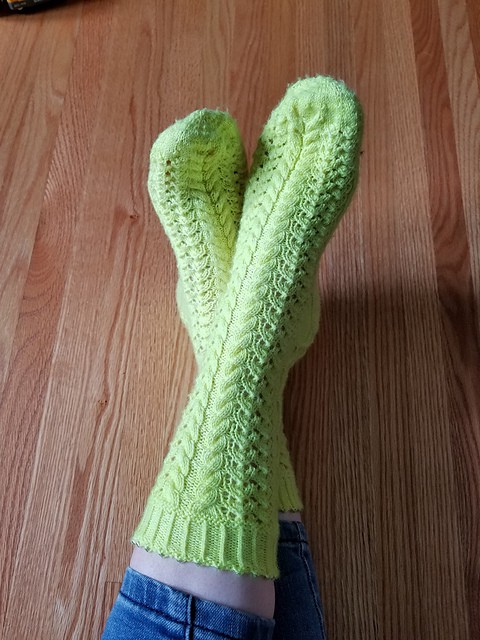 Feet wearing neon yellow lace and cable socks on a wood background
