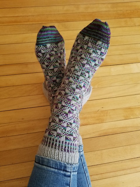 Feet wearing gray and purple variegated hand knit colorwork socks on wood background
